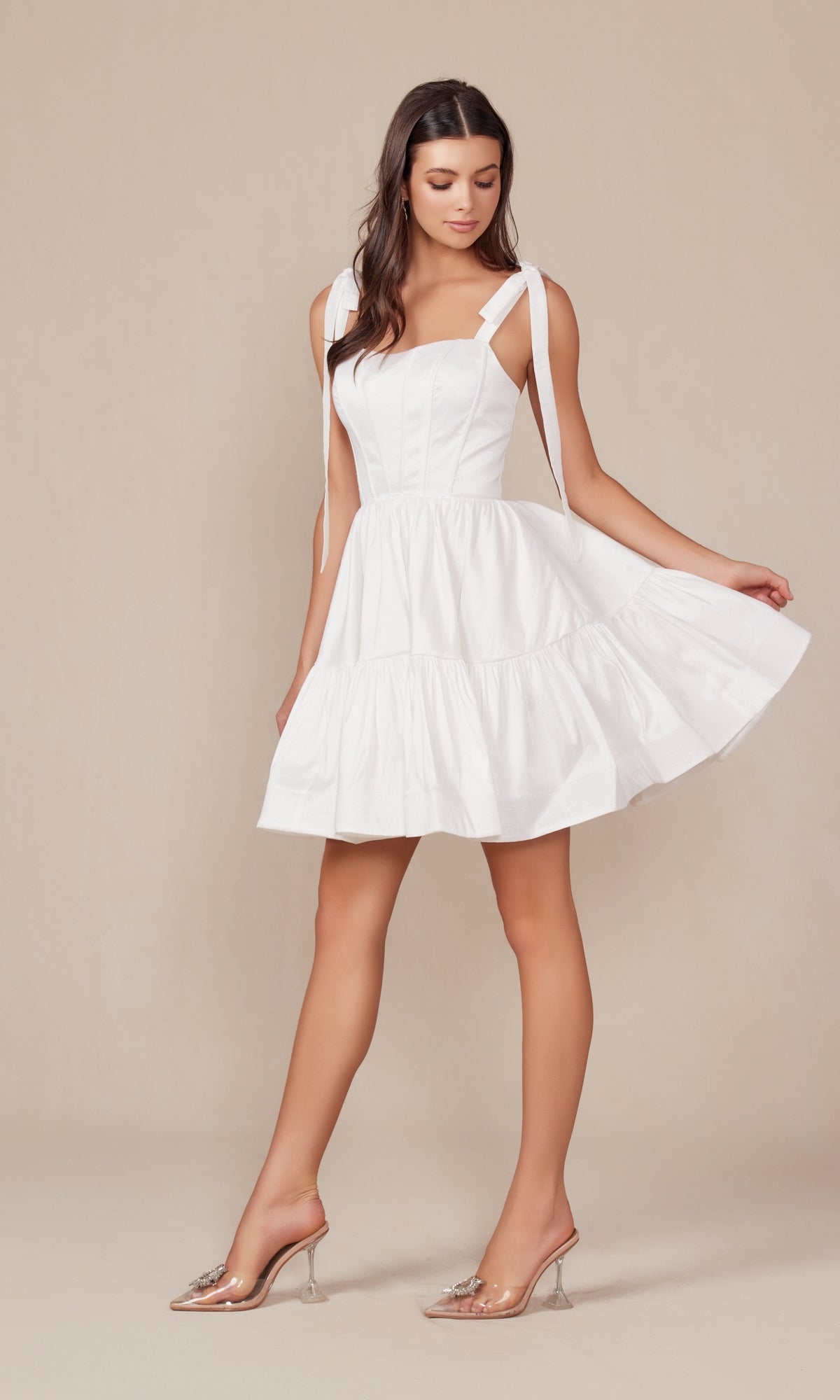 A woman stands against a plain background wearing the Shoulder-Tie Little White Party Dress T869W, featuring ribbon shoulder straps, a tiered design, and paired with clear high-heeled shoes.